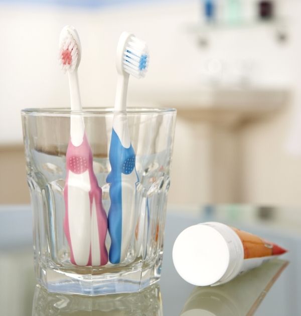 Eine rosa-weiße und eine blau-weiße Kinderzahnbürste in einem Glasbecher mit liegender Zahnpasta daneben