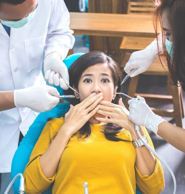 Frau mit Zahnarztangst auf dem Behandlungsstuhl, während das Personal mit Instrumenten ausgestattet mit der Behandlung beginnen will